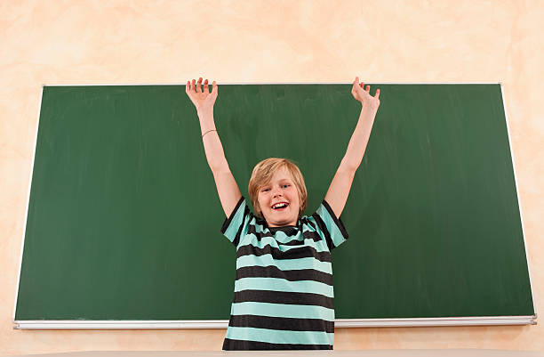 A funny young boy making glasses with his hands against a classroom blackboard