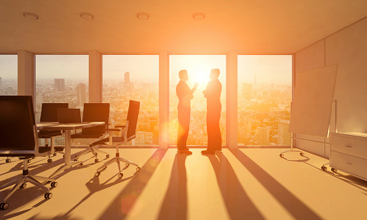 Silhouettes of two business men talking in front of a window inside a contemporary office with a conference desk, while warm sunset light fills the room creating lens flares. Outside the windows the skyline of a city with skyscrapers is visible. 