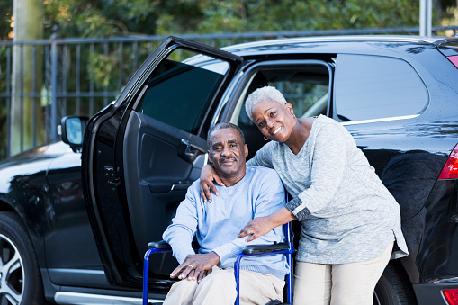 A disabled senior African American man has gotten out of a car, and is sitting in his wheelchair.  His loving wife is standing beside, with her arm around him.  They are smiling at the camera.