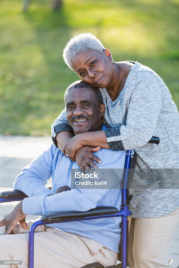 Senior African American couple, man in wheelchair Portrait of a senior African American couple outdoors.  The man is sitting in a wheelchair, smiling.  His devoted wife is standing behind him with her arms wrapped around his shoulders. Serious Stock Photo