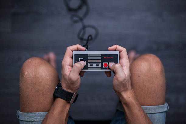 Nintendo Entertainment System - Video Game Controller Gothenburg, Sweden - January 31, 2015: A shot from above of a young man's hands using a Nintendo game controller, a remote controller for the Nintendo Entertainment System developed by Nintendo Co., Ltd. in the 1980s. Natural lighting. Shot on a grey wooden background. brand name games console stock pictures, royalty-free photos & images