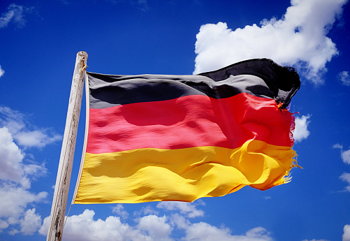 A rather tattered and frayed national flag of Germany billowing in the wind against a cloud-strewn sky.
