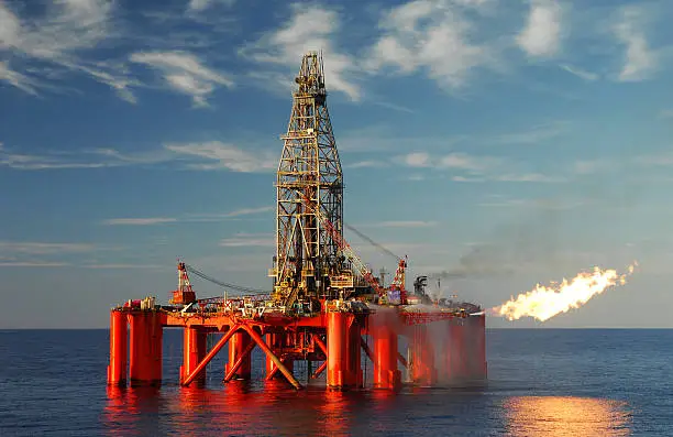 An oil rig situated in a calm blue ocean exploring for oil and gas. The oil rig is flaring from the side and this is reflected in the ocean. Fluffy white clouds are scattered in a blue sky.