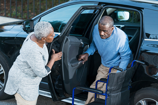 An African American senior woman helping her disabled husband climb out of a car into his wheelchair.