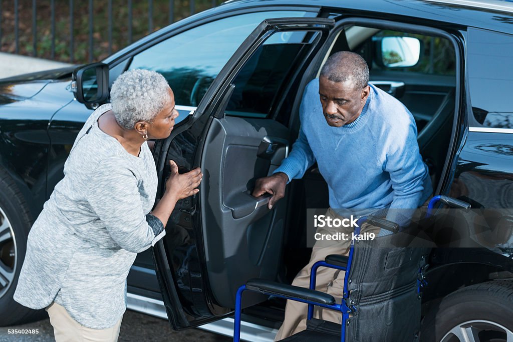 Senior woman helping disabled husband out of car An African American senior woman helping her disabled husband climb out of a car into his wheelchair. Car Stock Photo