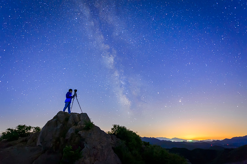 A photographer taking a photo of the night sky from the top of a mountain. 