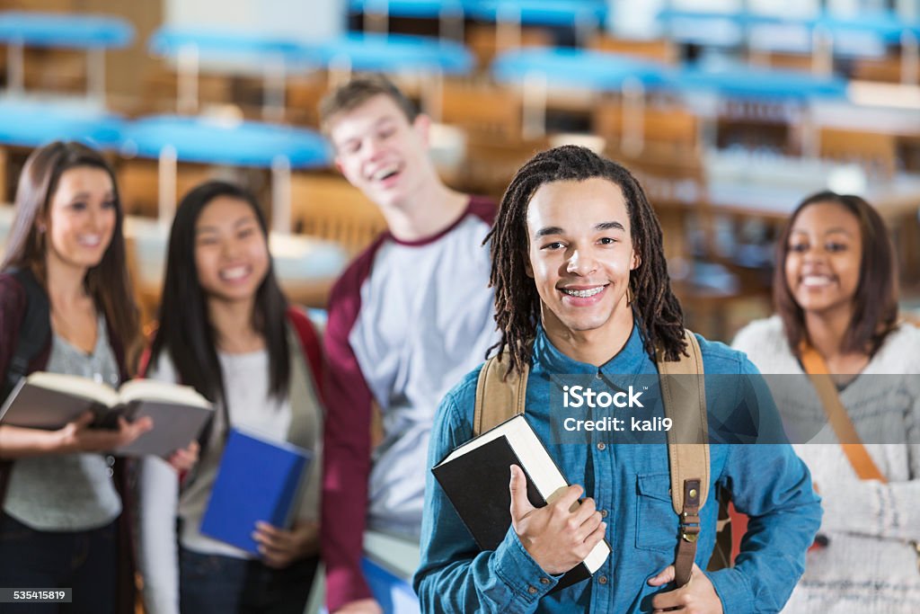 Group of students in library reading room A young man with dreadlocks is standing in the library with a textbook in his hand, smiling at the camera.  A group of students are out of focus behind him. 16-17 Years Stock Photo