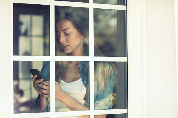 jovem mulher um utilizar um smartphone atrás de uma janela - mullion windows imagens e fotografias de stock