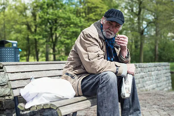 Photo of Homeless man smoking cigarette on bench in park.