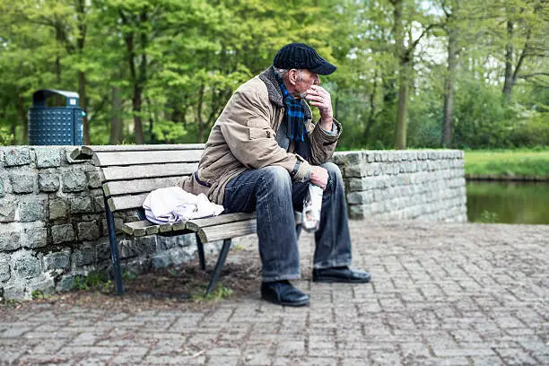 Photo of Smoking homeless man sitting on bench in park.