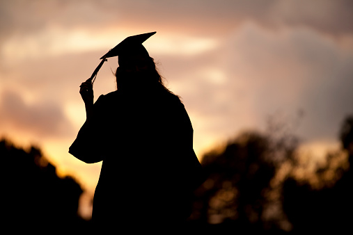 A female graduate is silhouetted by sunset in her cap and gown on a college campus