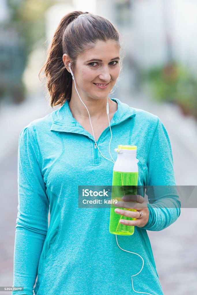 Woman with earbuds holding sports drink Mid adult woman listening to music through earbuds and holding a sports drink.  She is smiling at the camera. 2015 Stock Photo