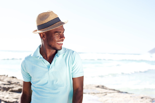 Portrait of stylish young african man with hat looking away and smiling at the beach in summer