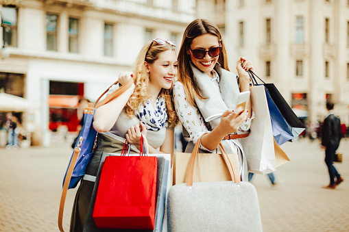 Cheerful beautiful women are shopping in the city. They are using smart phone, and one of them is holding a credit card.