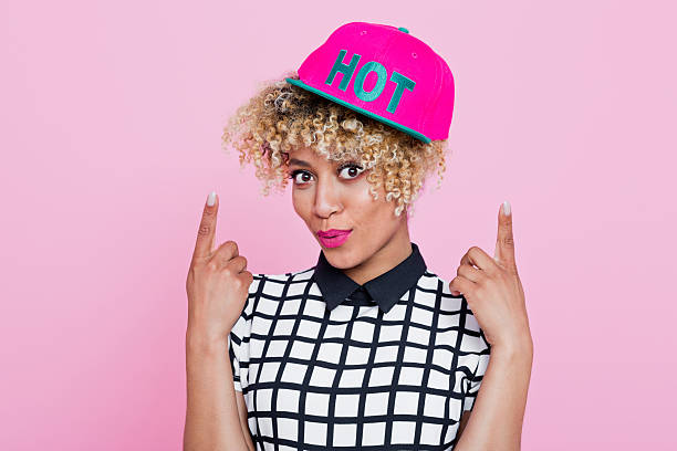 Summer portrait of happy, afro american young woman wearing grid check playsuit and pink baseball cap, standing with here fingers showing text HOT written at her cap, looking at camera and smiling. Studio shot, pink background.