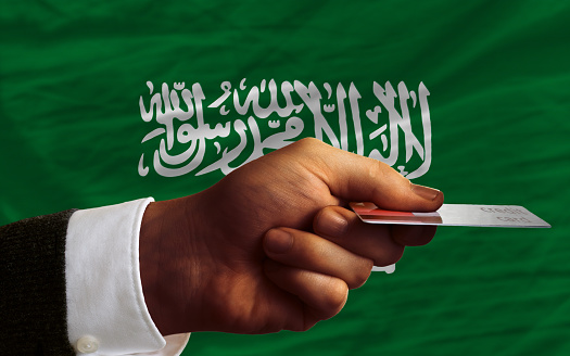 man stretching out credit card to buy goods in front of complete wavy national flag of saudi arabia