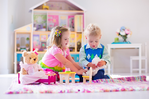 Kids playing with doll house and stuffed animal toys. Children sit on a pink rug in a play room at home or kindergarten. Toddler kid and baby with plush toy and dolls. Birthday party for little child.