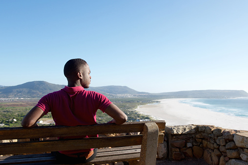 Rear view portrait of young african man sitting relaxed on a bench and looking at the sea, man on a holiday.