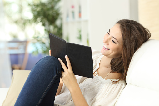 Relaxed happy woman reading a book in an ebook reader sitting on a couch at home