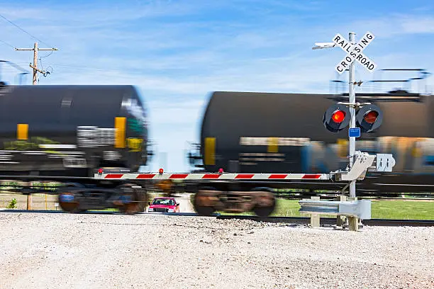 Railway level crossing on rural road with safety sign, flashing red lights and barrier as a tank train passes while SUV waits.  Long exposure with crossing sign in sharp focus and train behind with motion blur.  In camera motion blur, horizontal, copy space.