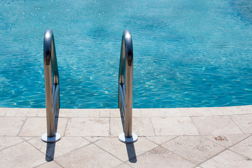 Image of the top of the shiny rails to a pool ladder.  The swimming pook decking is tile and the water appears blue, clean and refreshing.  The ladder rails are silver, clean and shiny.