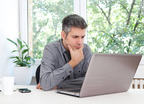 Working from home in a distraction-free space, a hairless, middle-aged man in his 50s appears to be making a video call on his laptop. He wears an earpiece in his ear.