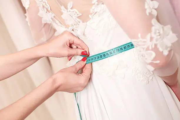Close up of arms of tailor measuring female waist with tape-measure. Young bride is trying on a wedding dress