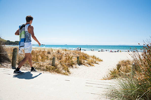 Mount Maunganui beach scenes. Mount Maunganui, New Zealand - January 22, 2015:  youth walks to beach on hot summer day, carrying his towel  at Mount Maunganui Mount, New Zealand. The beaches are a key summer destinations attracting domestic and international tourists mount maunganui stock pictures, royalty-free photos & images
