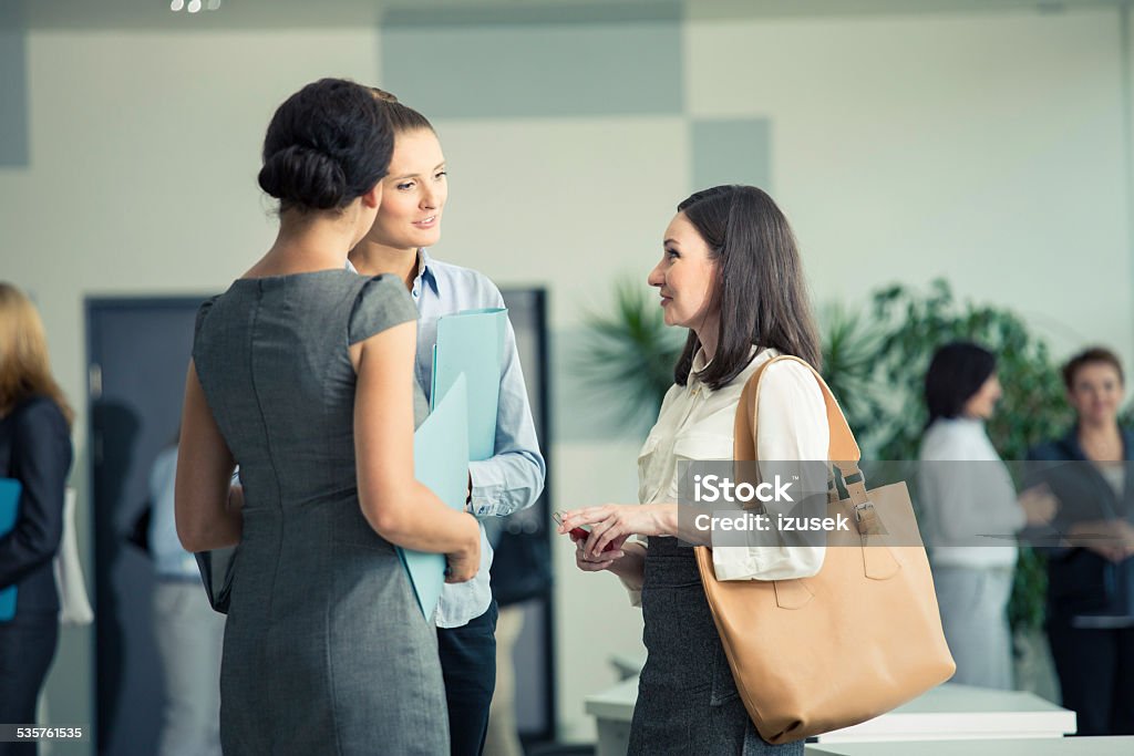 Three businesswomen talking in an office Three businesswomen standing in the circle in a modern office hall and talking together. People in the background. 2015 Stock Photo