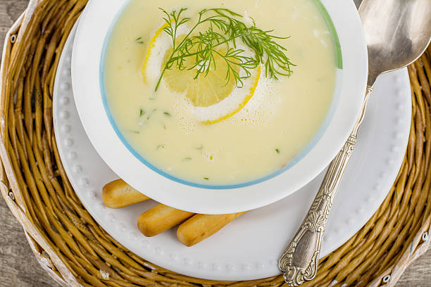 Greek soup avgolemono with a circle of lemon on a white plate on a wicker tray and wooden background with German silver spoon and bread sticks