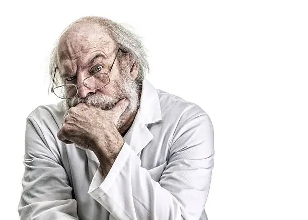A bizarre, skeptical, messy, brilliant, genius, tangled haired psychiatrist (or he might be a poorly groomed doctor, scientist, pharmacist, or some other medical occupation professional) is looking at the camera while listening closely with a serious, raised eyebrow facial expression. He is a balding senior adult man with a gray beard and mustache. He is wearing a white lab coat and is sitting with left hand covering his chin. Canon 5D Mark III.