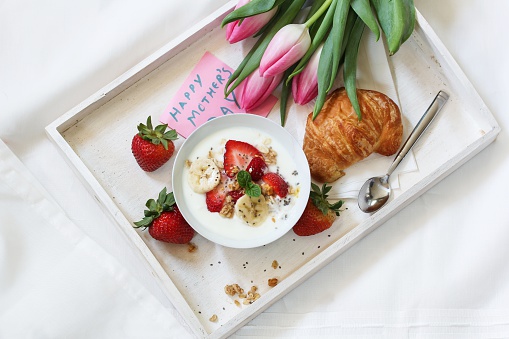 Breakfast yogurt bowl with strawberries and bananas served in a white tray with croissant Mother's day note and tulip flowers / Mother's day Brunch