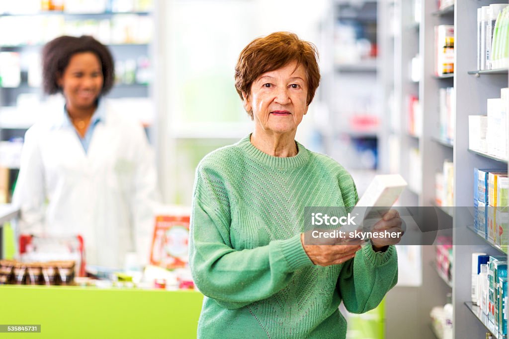 Female customer in a pharmacy. Senior woman in a pharmacy choosing a medicine to buy and looking at the camera. African American pharmacist is in the background.   Customer Stock Photo