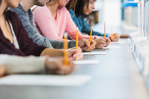 Cropped view of a multiracial group of young men and women sitting in a row at a table, writing with pencils on paper.  They are taking a test or filling out an application.  Focus is on the hand of the young man in the middle in the gray shirt.