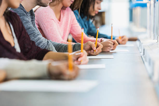 Cropped view of group of teenagers taking a test Cropped view of a multiracial group of young men and women sitting in a row at a table, writing with pencils on paper. They are taking a test or filling out an application. Focus is on the hand of the young man in the middle in the gray shirt. exam paper stock pictures, royalty-free photos & images