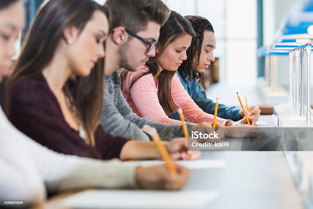 Group of teenagers taking a test Side view of a multiracial group of young men and women sitting in a row at a table, writing with pencils on paper.  They are taking a test or filling out an application.  Focus is on the girl in the pink shirt. Educational Exam Stock Photo