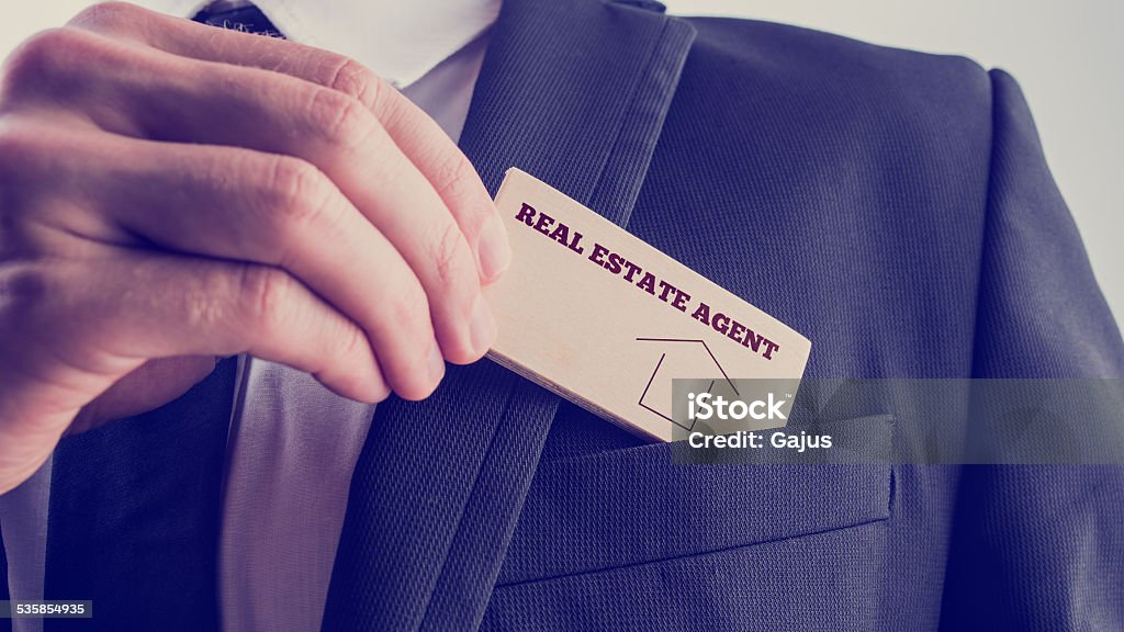 Agent in Simple Real Estate Concept Real Estate Agent in Black Suit Putting Small Wooden Piece with Real Estate Agent Text and Graphic to Front Pocket. 2015 Stock Photo