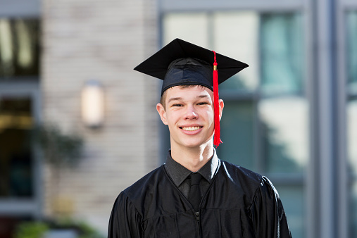 Portrait of a teenage boy at his high school graduation, wearing a black cap and gown with a red tassel.