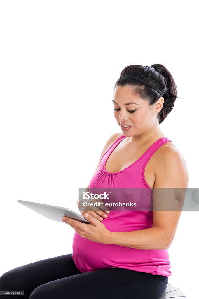 Fit pregnant woman looking at tablet PC Beautiful mixed race Hispanic woman 7 months pregnant sitting on an exercise ball looking at tablet pc isolated on white background Athlete Stock Photo