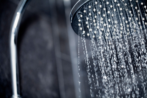 Close up view of large shower head sprinkling water with black tiles in the background.