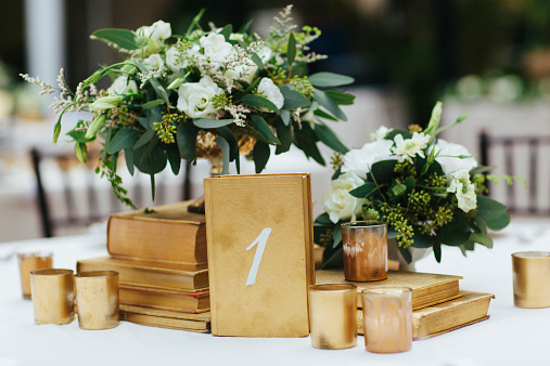Close up photograph of elegant decor on a table at a wedding reception. Gold accents include the number one and flower arrangements have a green and white color theme.