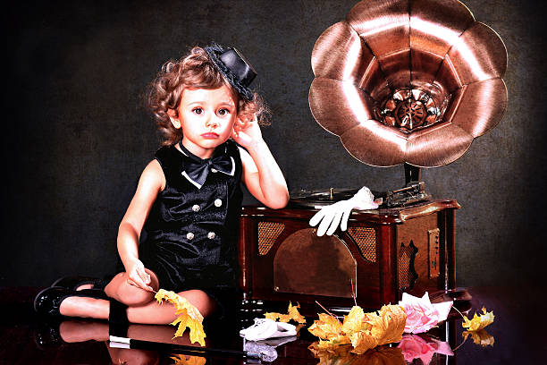 Cute baby in stylish black dress and hat listening to music on the gramophone with her hand to her ear