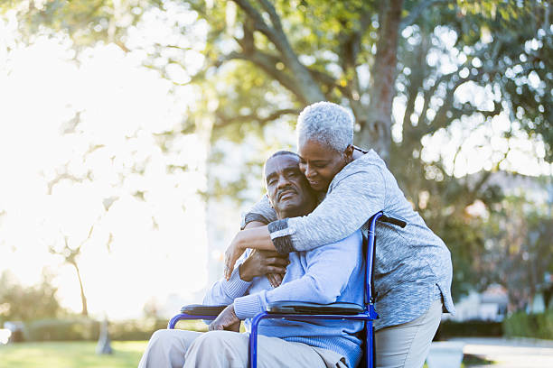 Senior African American couple, man in wheelchair Portrait of a senior African American couple outdoors, showing their affection in the bright sunlight.  The man is sitting in a wheelchair, in the warm embrace of his devoted wife.  Their eyes are closed. stroke illness stock pictures, royalty-free photos & images