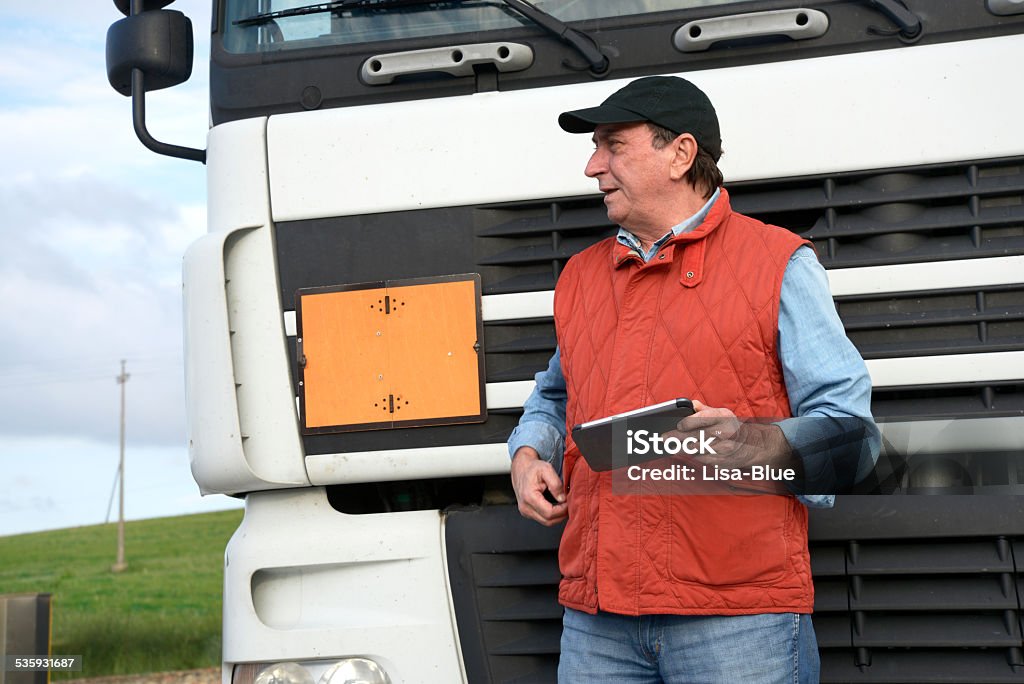 Truck Driver Truck driver standing with his back in front of his truck. He is looking on the left and holding a tablet. In the background a gas station and a green field.  2015 Stock Photo