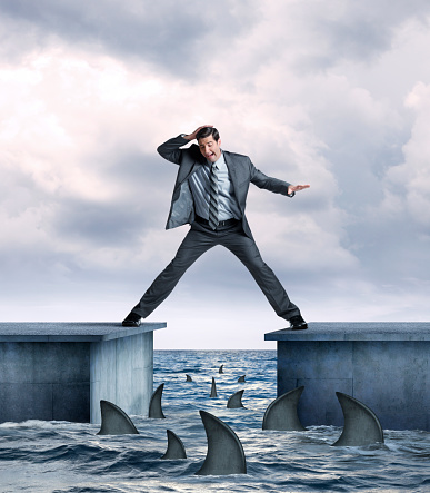 A businessman straddling two piers  tries not to fall into shark infested  ocean waters below.  Several sharks are circling the pier as the businessman's legs are stretched as far as they can go as he balances himself.  His hand is on top of his head as he looks down at the sharks below in fear.  