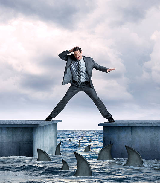 Businessman In Danger Of Falling Into Shark Infested Waters A businessman straddling two piers  tries not to fall into shark infested  ocean waters below.  Several sharks are circling the pier as the businessman's legs are stretched as far as they can go as he balances himself.  His hand is on top of his head as he looks down at the sharks below in fear.   legs apart stock pictures, royalty-free photos & images