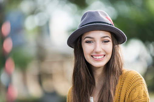 Face of a beautiful teenage girl with long, straight, brown hair smiling at the camera.  She is standing outdoors, wearing a sweater and brimmed hat.