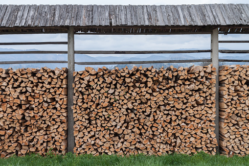 Pile of wood stacked outdoors on a farm near Bled, Slovenia. The wood is cut to uniform size and stacked in a neat row under a shelter. The shelter is made up of tall posts and a short, sloping, wooden roof. Foreground is green grass and mountains in the distance. 