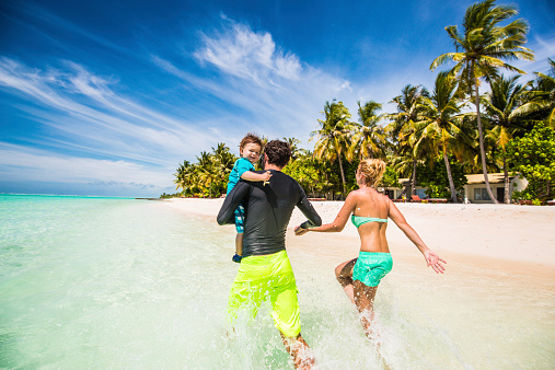 Young happy family enjoy their vacation by the seaside. They are jogging together through the crystally clear water of the Maldivian islands, while holding hands.