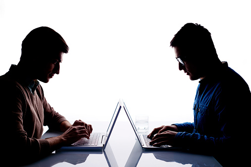 Back lit image of two men sitting at table in front of each other and typing on laptops